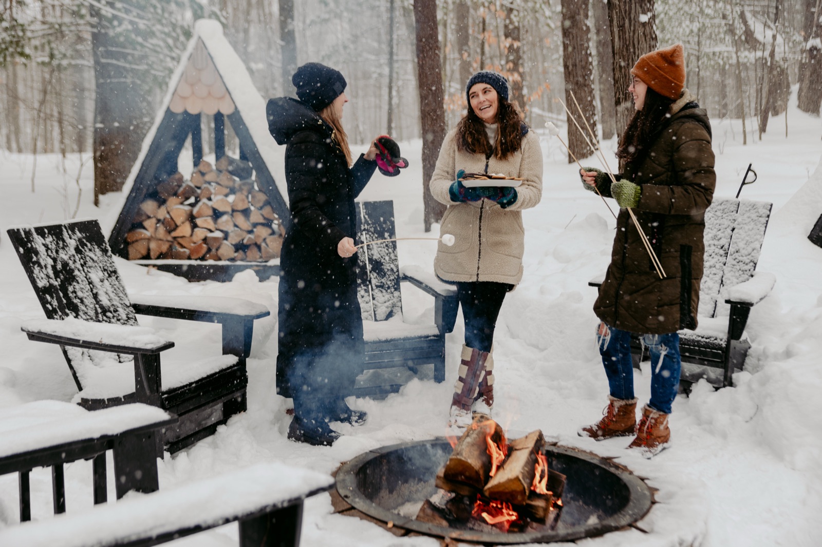 Three guests toasting marshmallows around a crackling fire pit in falling snow in front of the Roost A-Frame cabin near Lake Ann