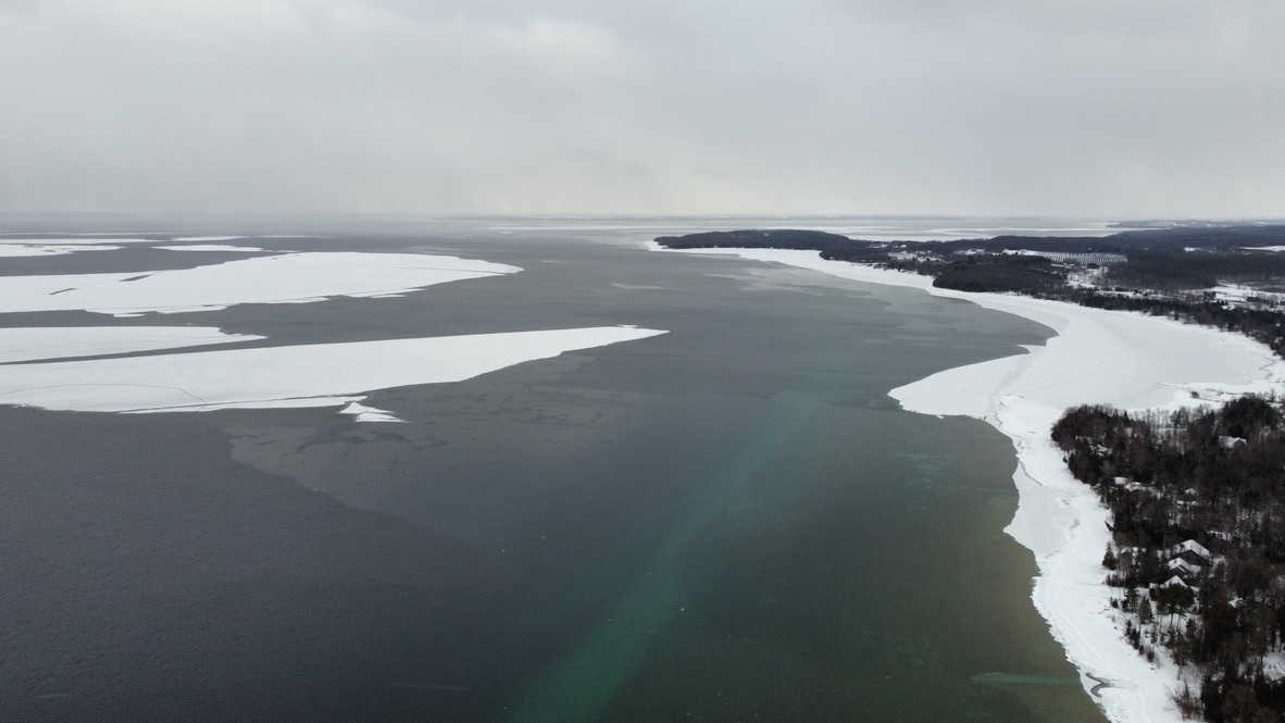 Aerial view of Grand Traverse Bay in winter with dark open water meeting white ice along a snow-covered forested shoreline