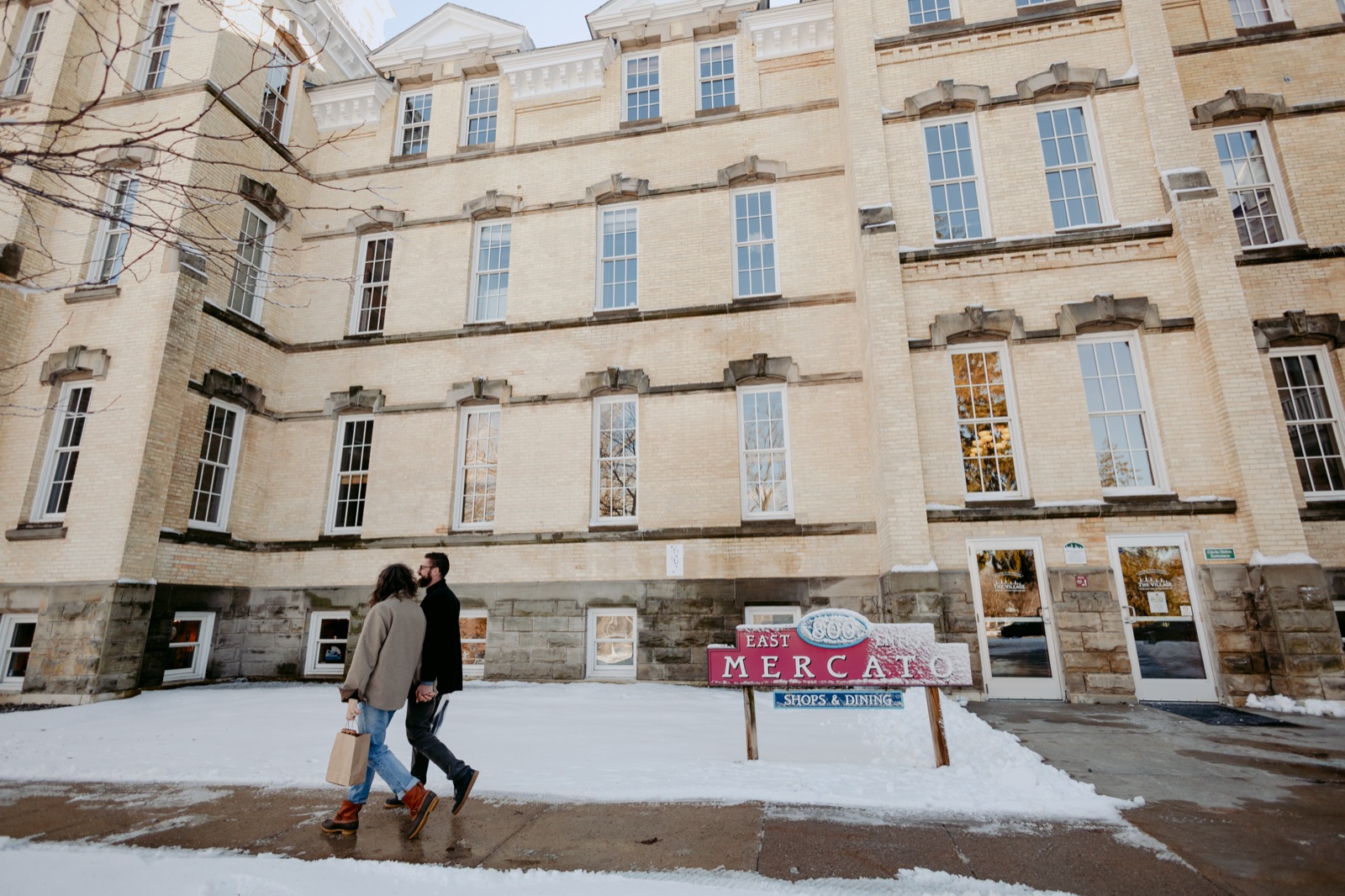 Couple walking hand in hand past the historic brick buildings of the snowy Grand Traverse Commons in Traverse City