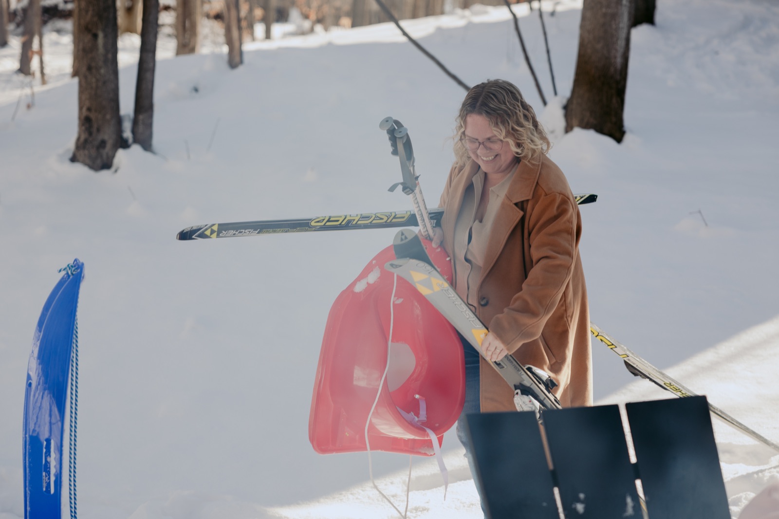 Smiling woman in a camel coat carrying cross-country skis and a sled in the snowy woods near Traverse City